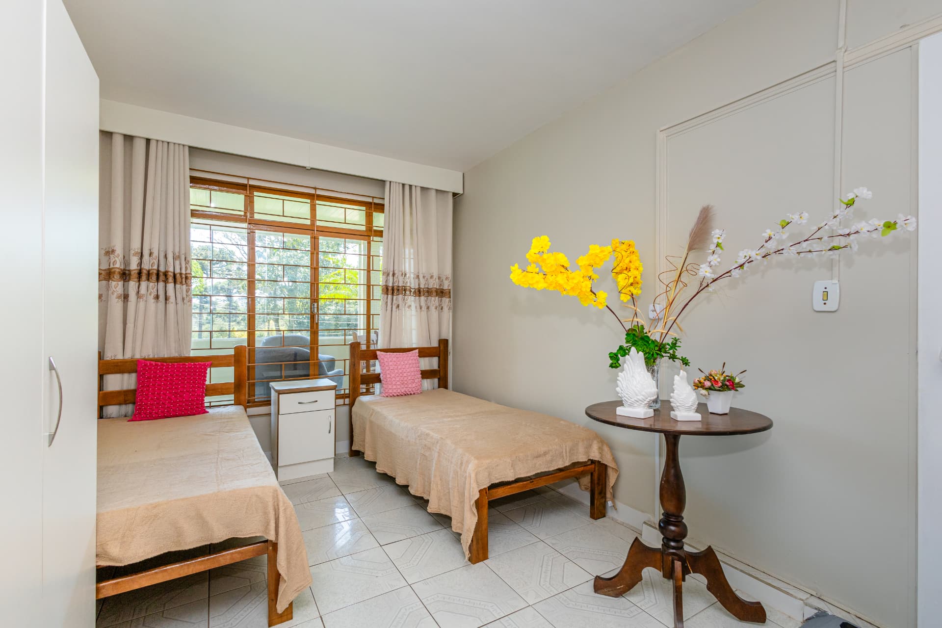 Quarto com duas camas, criado-mudo branco e mesa com flores